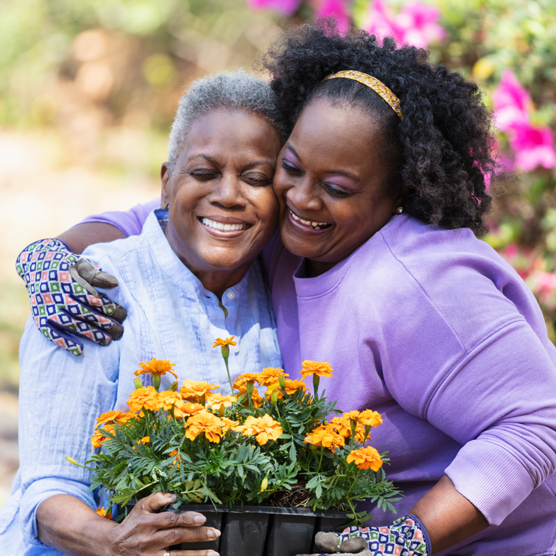 A caregiver providing companion services for seniors hugs an older adult while they both hold a flower pot filled with yellow flowers.