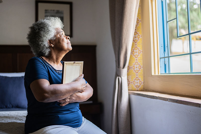 A woman looks thoughtfully out of a window while holding a framed photo. Grief in older adults is complex and requires support.