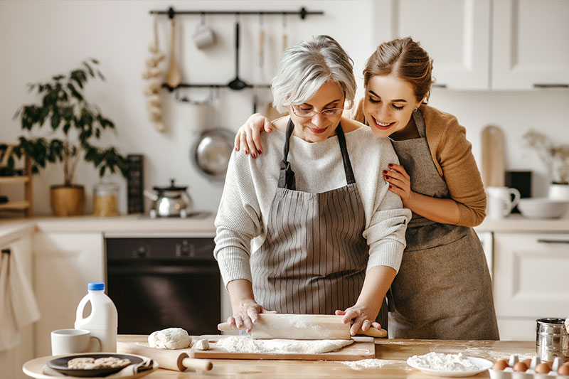 A woman looks on approvingly as her mother is rolling out dough in the kitchen. She knows that cooking together is a therapeutic tool for seniors with dementia.