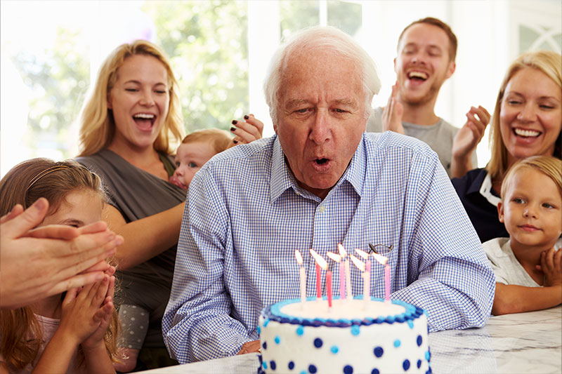 An older man’s family, who spent time researching birthday celebrations for seniors, smile as they watch an older loved one blow out the candles on his cake.
