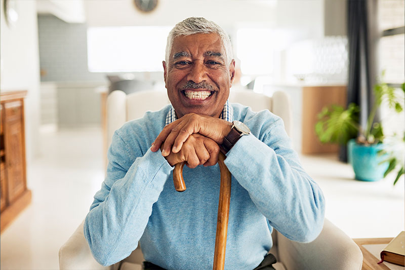 A man smiles while holding a cane at his home. Addressing the fear of falling in older adults promotes independence. 